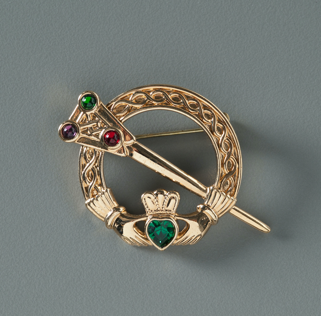 A gold Irish Claddagh brooch belonging to Kathleen Moran is displayed on a gray surface. The jewelry includes emerald stones. 