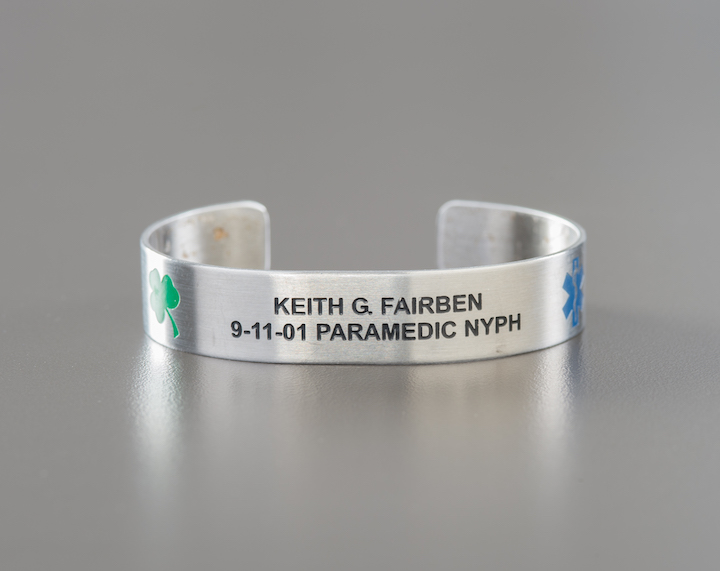 A metal memorial bracelet honoring Keith George Fairben. The bracelet features Fairben's name, as well as a green shamrock and a blue "star of life" paramedics symbol. 