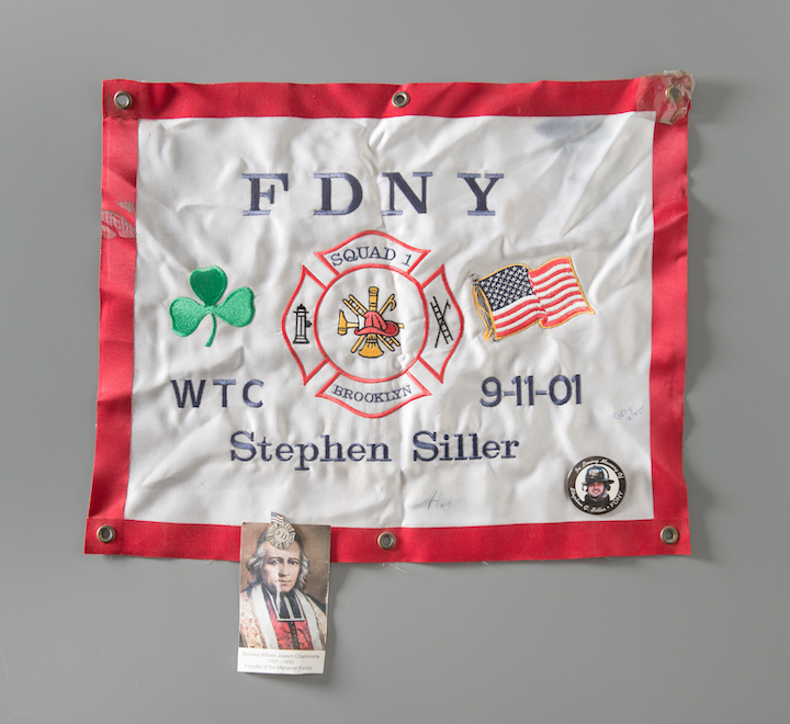 A white memorial flag honoring Stephen Gerard Siller is displayed on a gray surface. The flag has a red border and the letters FDNY printed across the top of it. A shamrock and American flag border a Maltese cross at the center of the flag. .  On the left is a card for Blessed William Joseph Chaminade attached with a lapel pin.  