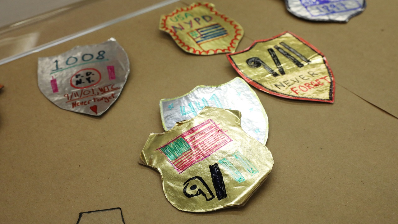Children's craft projects show six first responder badges made of foil and paper. In the foreground, a badge made of gold foil shows an American flag with the date nine eleven written below it. 