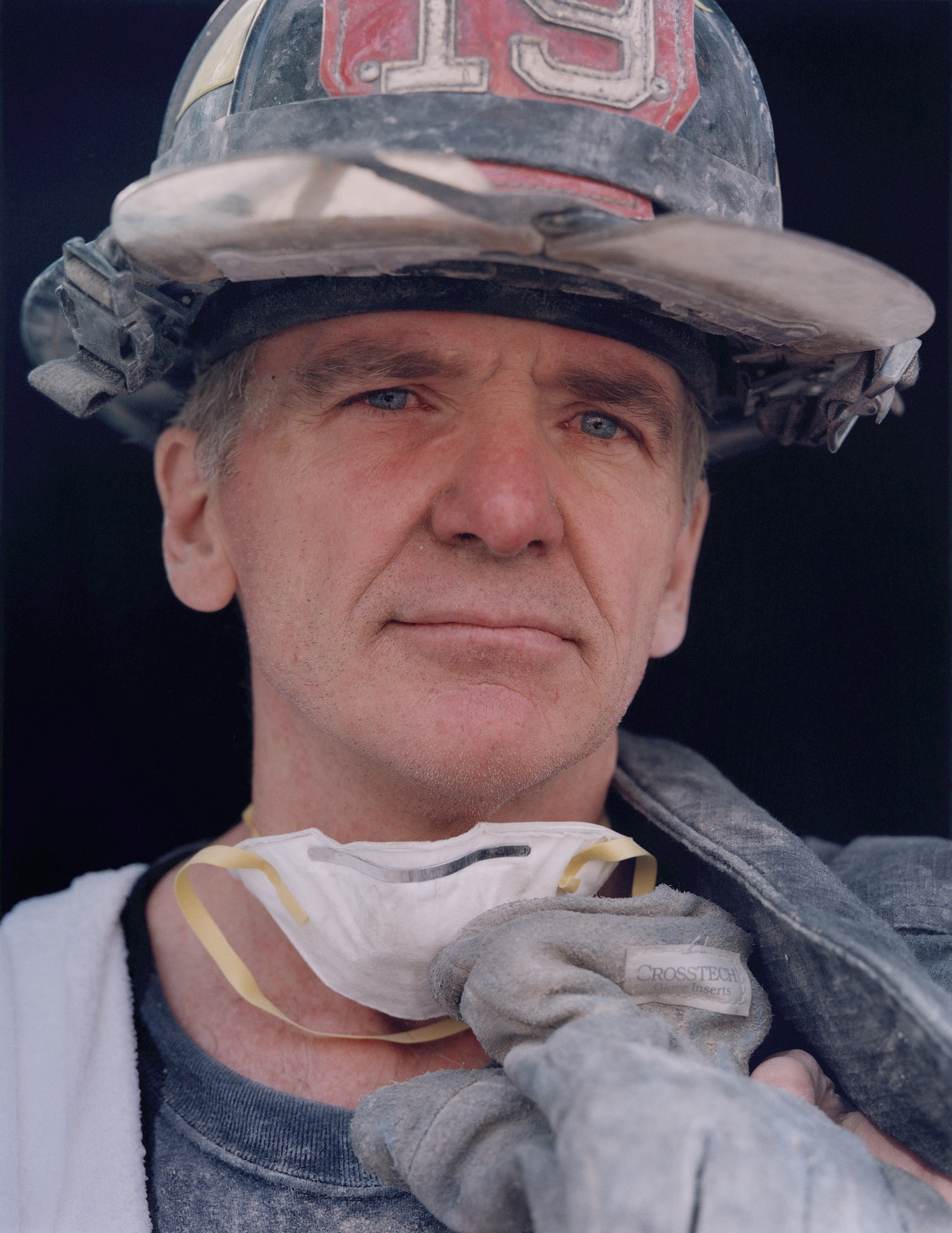 This photograph shows a man with a dust-covered fire helmet and a dust must around his neck. He looks directly into the camera. 