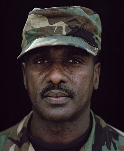 A man in a camouflage hat and jacket stares solemnly ahead. 
