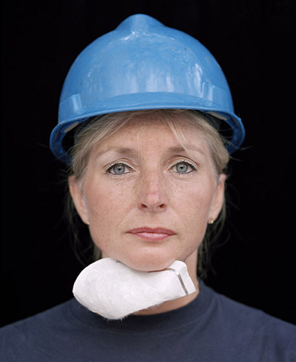 A woman in a light blue hardhat and a breathing mask worn on her neck  stares straight ahead in this photographic portrait. 