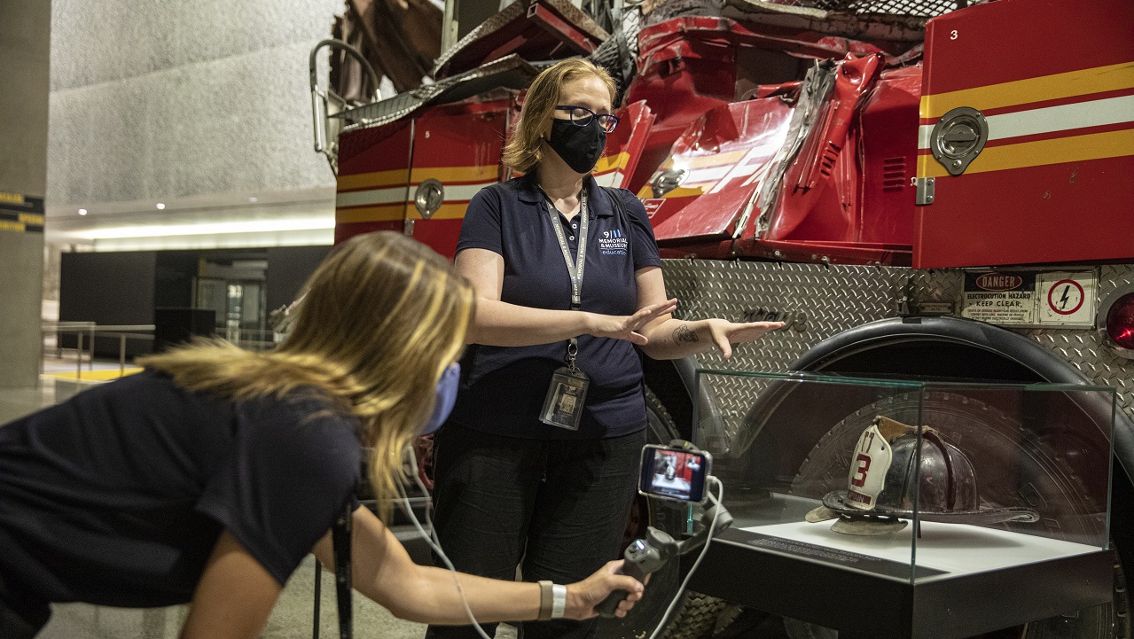 A woman wearing a face mask stands in front of the Ladder 3 firetruck in the Memorial Museum. Another woman films the scene with her smartphone. 