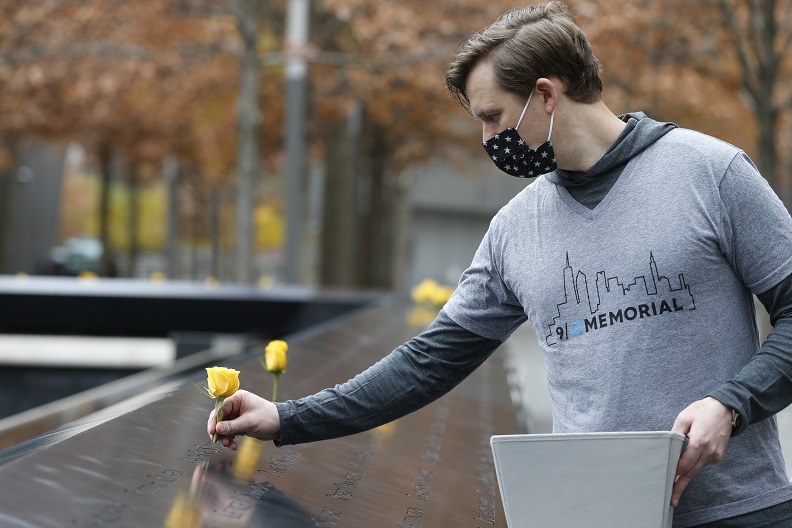 A man wearing a facemask and a gray 9/11 Memorial t-shirt places yellow roses into the bronze parapets. 