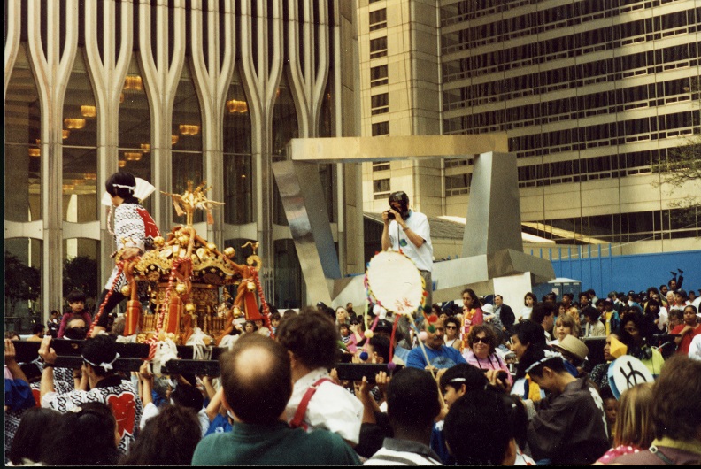 A large crowd of people in front of a Twin Tower. A man stands in front of an abstract steel sculpture of four intersecting beams taking a photograph of a procession carrying a pagoda-like object in red and gold. 