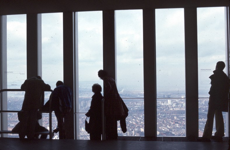 Silhouette shot of five visitors in front of a row of six full length windows, looking down to a bird's eye view of the city on a cloudy day. 