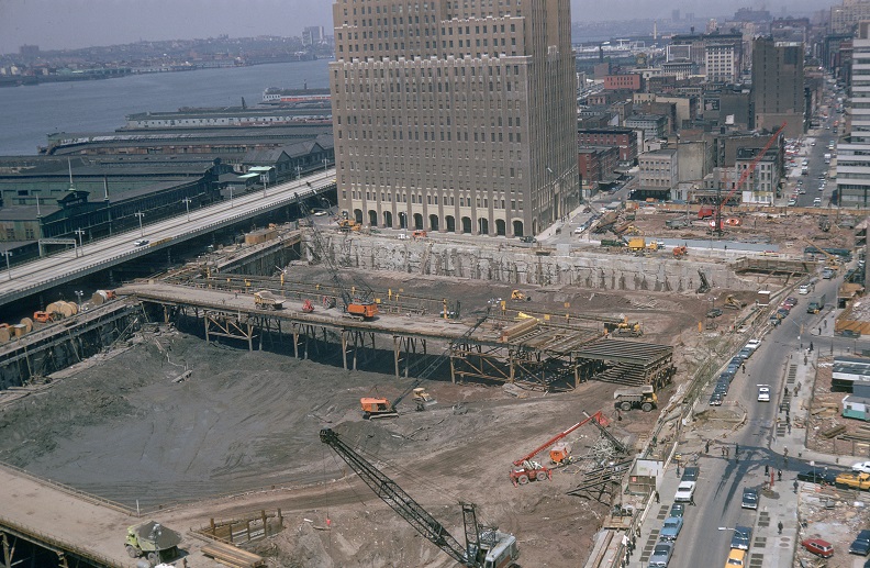Aerial view of a square excavation site in the foreground and buildings in the background with the Hudson river visible on the upper-left corner. 