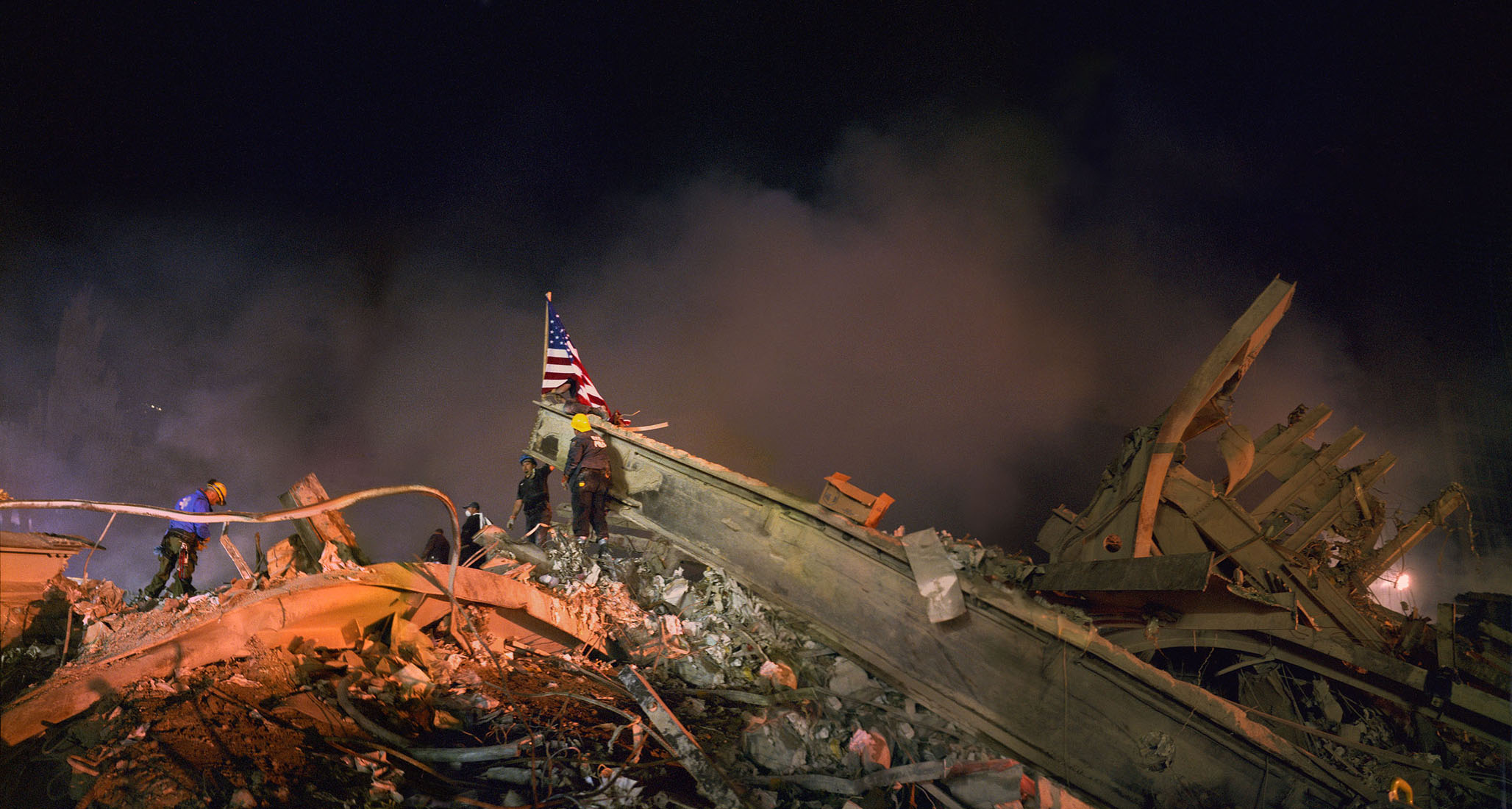Workers at Ground Zero placing a United States flag atop a steel beam that lays atop the pile. Photo taken at night.