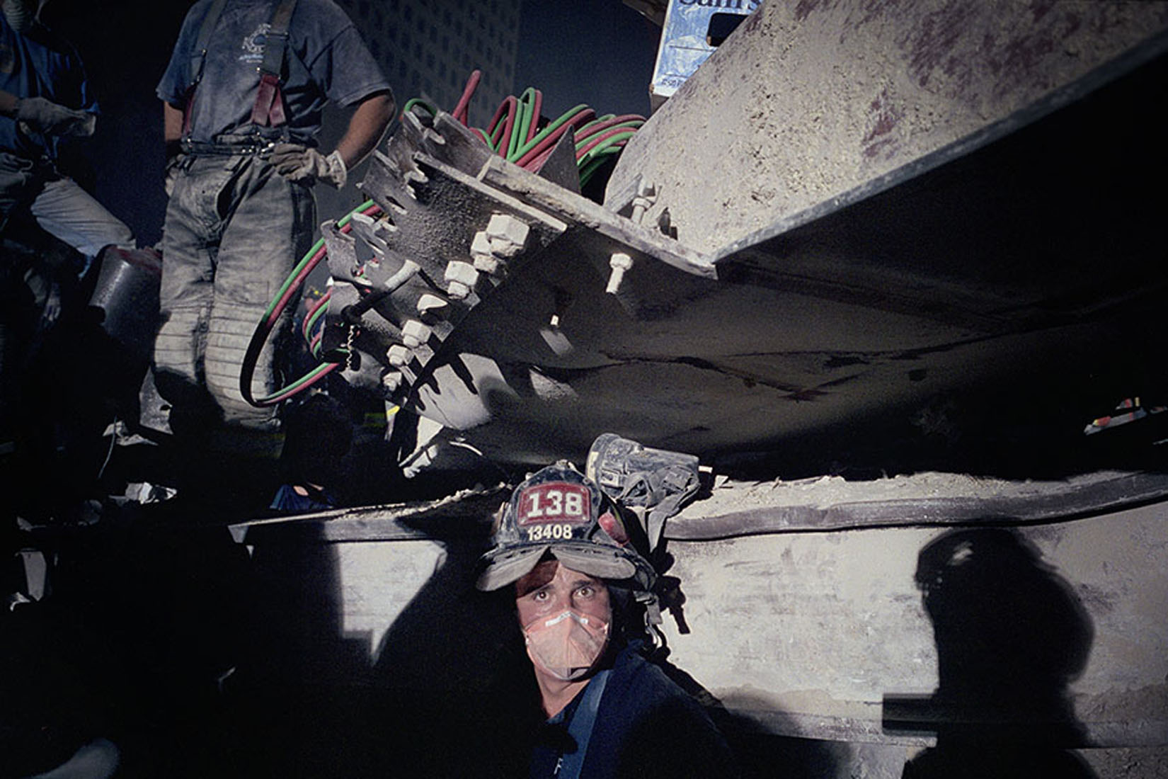 A firefighter at Ground Zero looks at the photographer from underneath an ash-covered metal beam. Firefighter is wearing helmet 138 and a surgical mask.