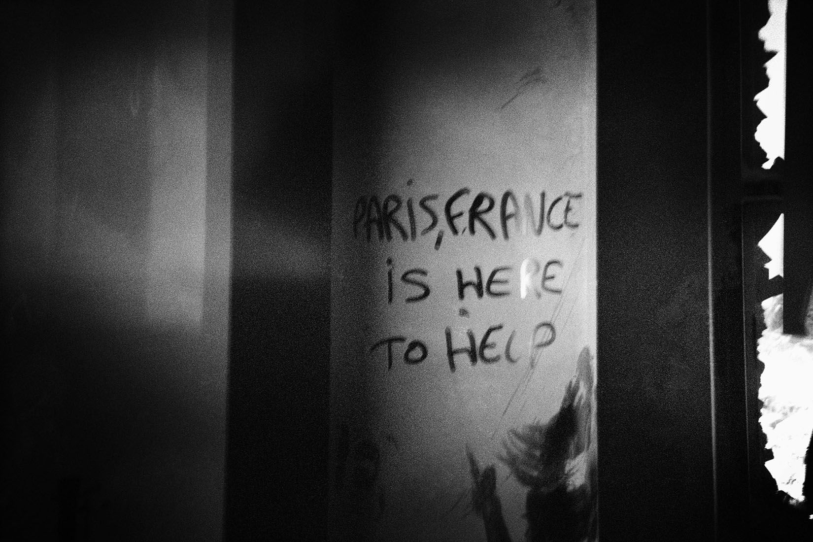 Black and white photo of an ash-covered surface. Someone has used a finger in the dirt to write Paris, France is here to help.