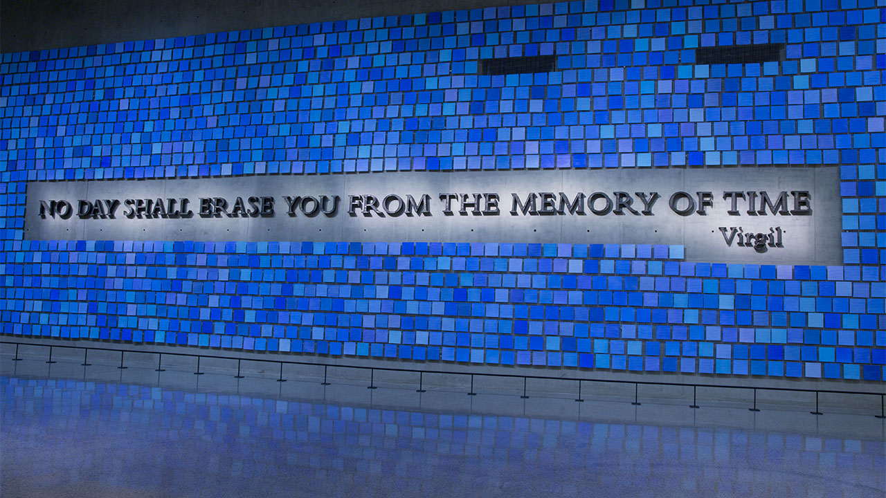 A wall of tiles, each a different shade of blue, with the inscription "No day shall erase you from the memory of time" across it.