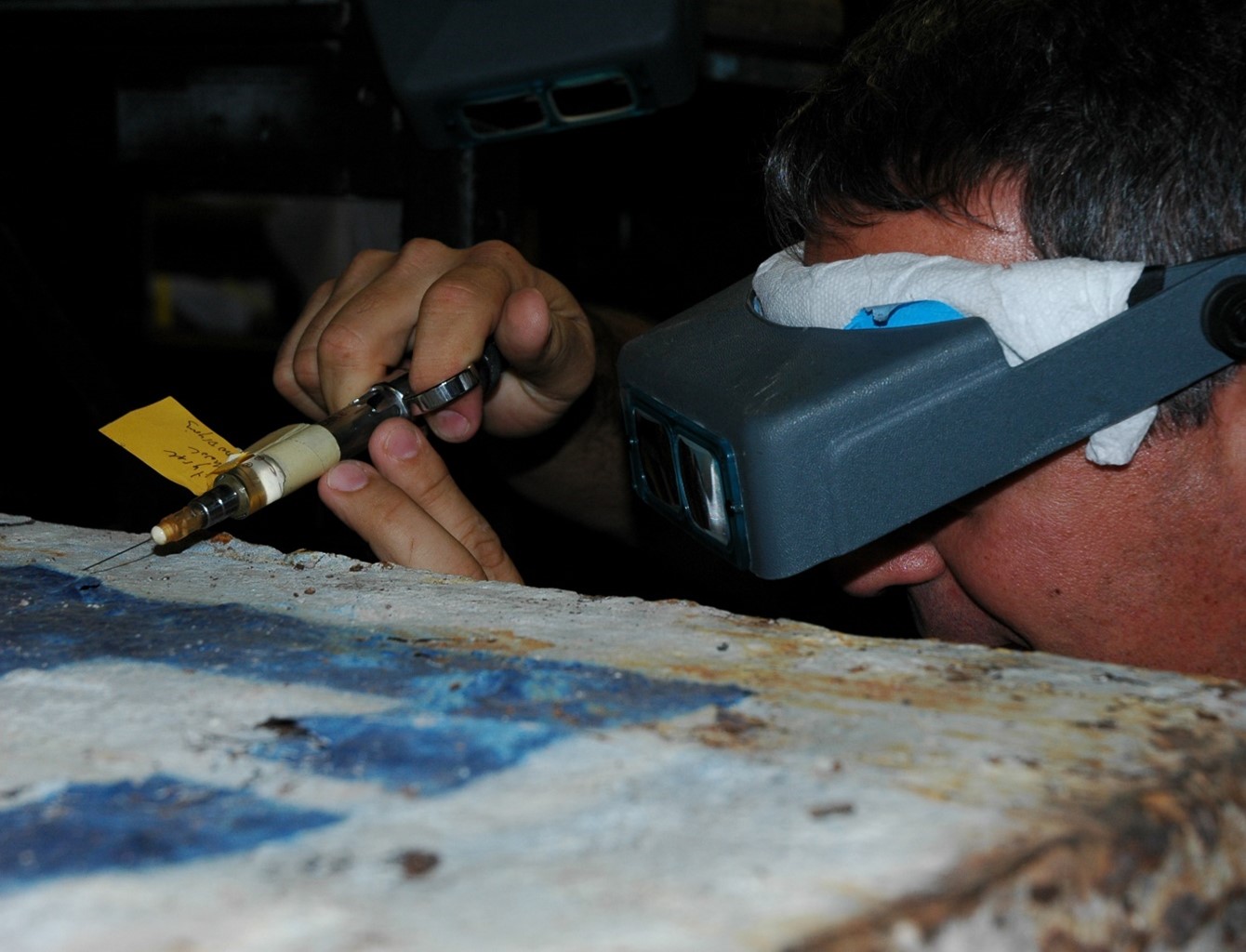 Man with eye equipment, including a paper towel wrapped inside the digital goggles, uses a syringe on an artifact of metal, which is rusted and painted