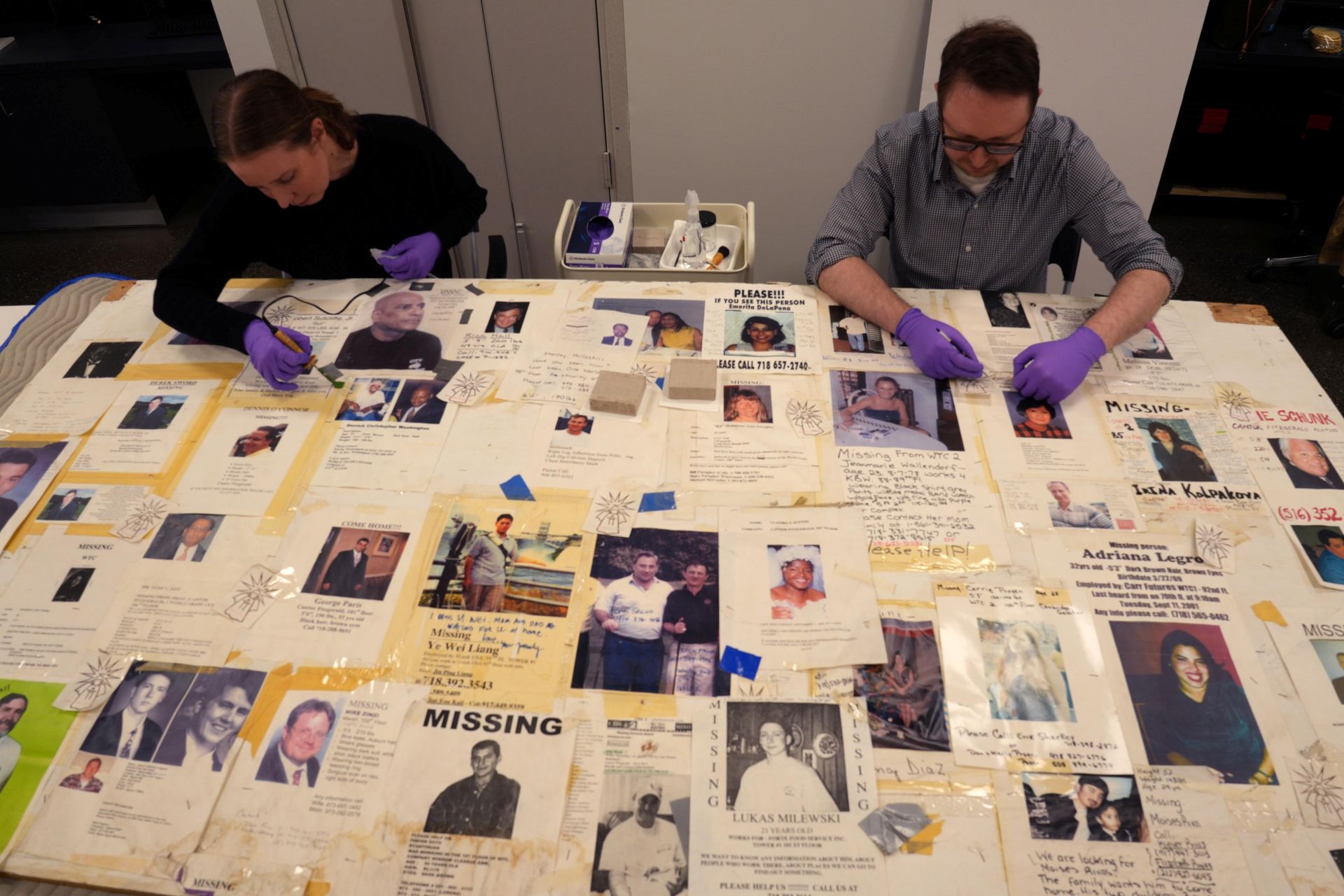 Two conservationists sit at a large table covered in missing persons posters, treating the different parts.