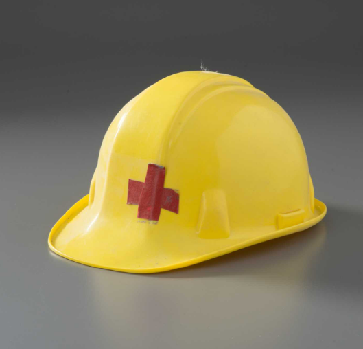 Plastic hard hat with cap-style brim and interior suspension. The hard hat is yellow with a red cross on the front created with duct tape. The hard hat is slightly dusty.