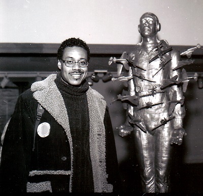 Black and white photo of a man standing and smiling in front of metallic sculpture of a stoic male with multiple small planes piercing on the body like arrows.