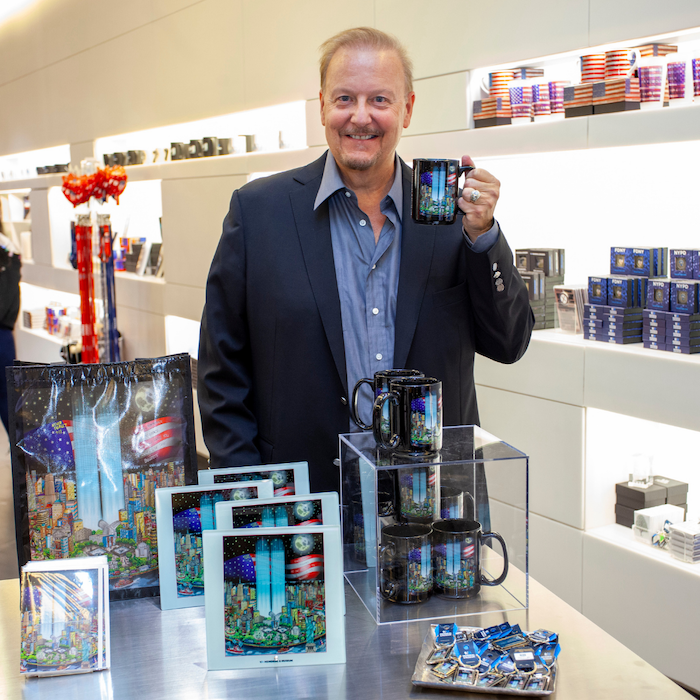 A bearded man in a dark jacket and shirt stands in front of a gift shop display featuring prints of his artwork, which depicts the Twin Towers, and mugs that feature the same design