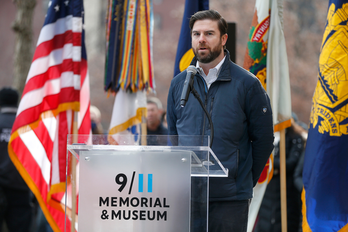 Bearded man in blue winter jacket stands at the 9/11 Memorial & Museum podium, with American flag on left