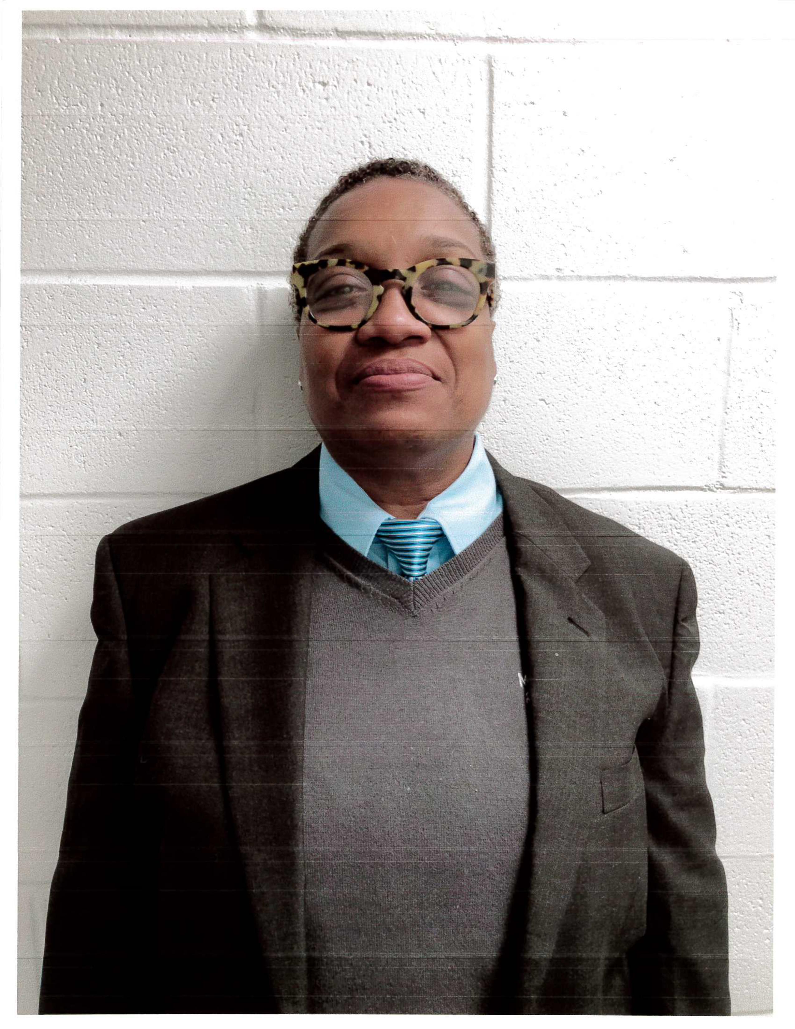 A woman with dark, cropped hair and black glasses stands against a cement wall wearing a light gray sweater, dark gray jacket, and blue blouse