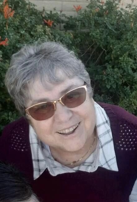 A woman with short gray hair wearing sunglasses and a plaid shirt under a burgundy sweater smiles up at the camera