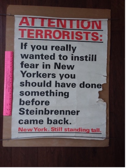A slightly battered poster with the text ATTENTION TERRORISTS in red, and IF YOU REALLY WANTED TO INSTILL FEAR IN NEW YORKERS, YOU SHOULD HAVE DONE SOMETHING BEFORE STEINBRENNER CAME BACK in black, with NEW YORK. STILL STANDING TALL in red at the bottom. 