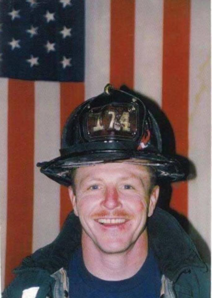 A man in a firefighter's helmet and uniform stands, smiling, in front of a vertical American flag
