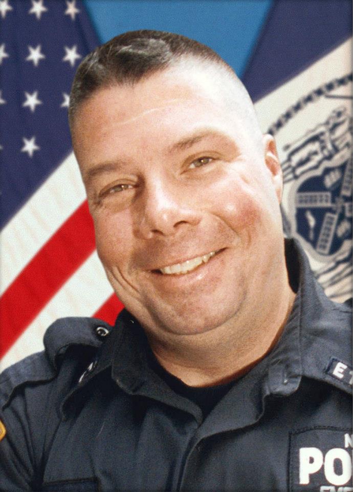 A smiling man with short, dark hair stands in front of an American flag in an NYPD uniform