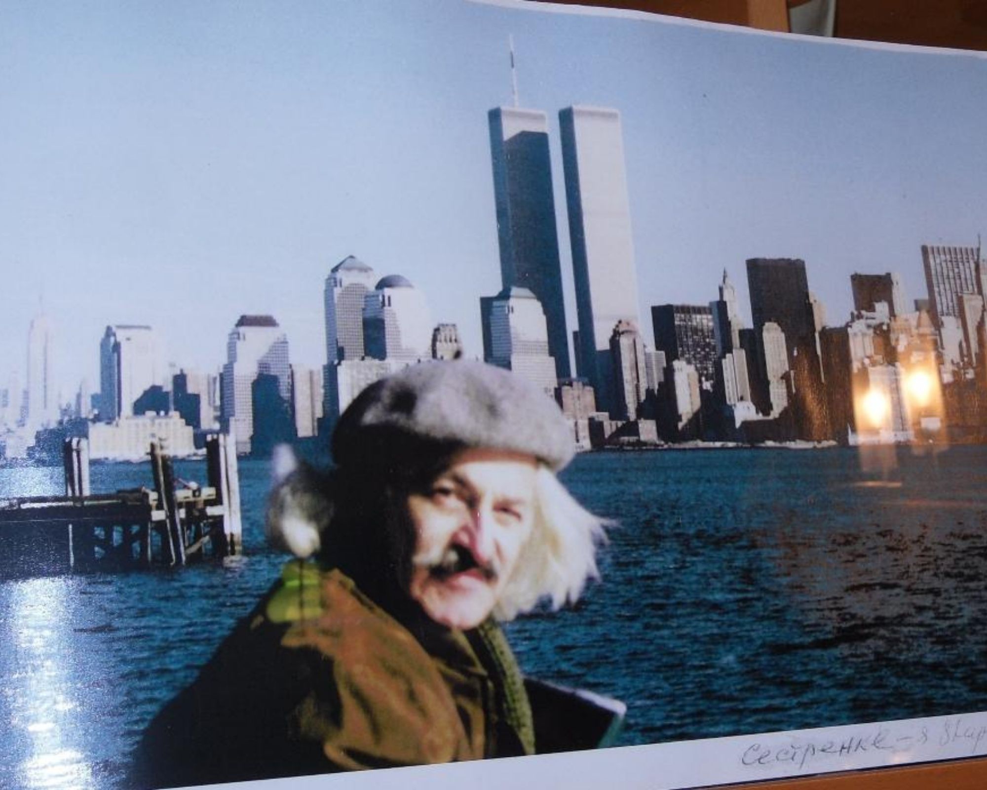 A gray-haired and mustached man with a brown jacket and gray cap poses in front of the lower Manhattan skyline, with the Twin Towers visible in the forefront