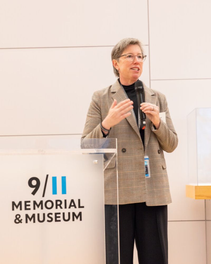Beth Hillman, President and CEO of the 9/11 Memorial & Museum, speaks at a glass podium in the Museum. The Museum logo appears on the podium and Beth is holding a microphone.