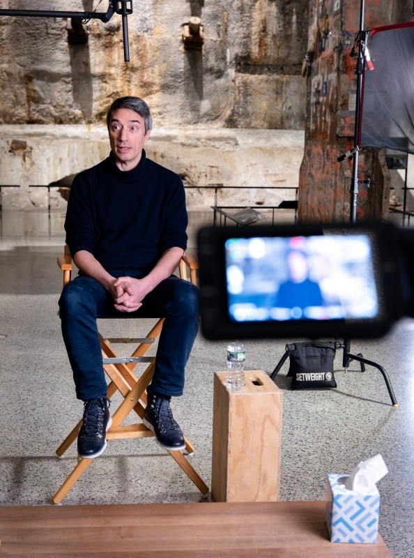 a man in a director's chair is being filmed, with the lens in the foreground