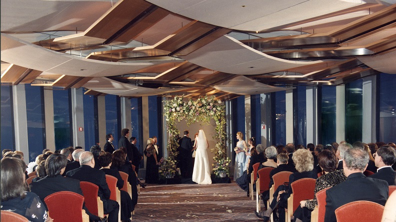 Bride and groom stand at the end of the aisle, facing towards a decorated background. Rows of guests seated on coral-colored velvet chairs on both sides of the aisle. 