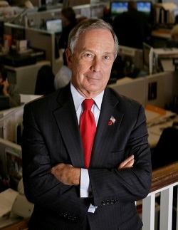 Michael R. Bloomberg, the chairman of the 9/11 Memorial Museum, smiles softly with his arms folded.  He is wearing a red tie and navy blazer. 