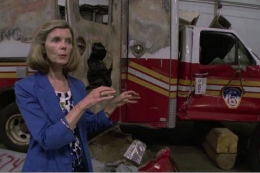 Jan Ramirez stands in the foreground wearing a bright blue blazer, gesturing in front of an FDNY truck