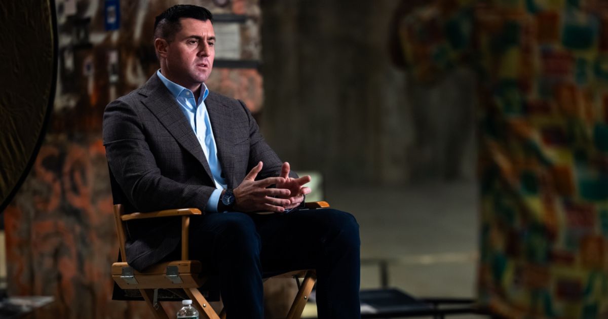 A man sits in a director's chair, with graffiti-covered concrete behind him