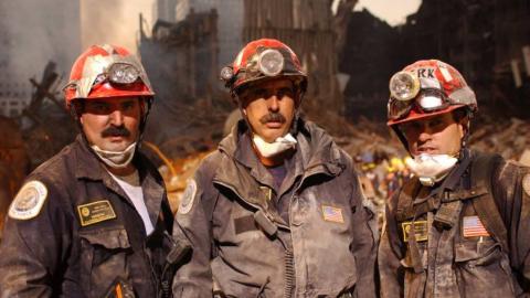 Three first responders wearing red hard hats and protective gear, and covered in dust, stand at Ground Zero