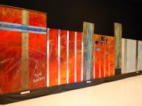 Long interconnected work with (l. to r.) Large wooden beam with crossing Greek key motif surrounded by brilliant red;  fiery red canvas panels with smaller forms, overlain by shiny metal laths; and two large abstract sections with white-obscured sky blue, overlain with metal laths.