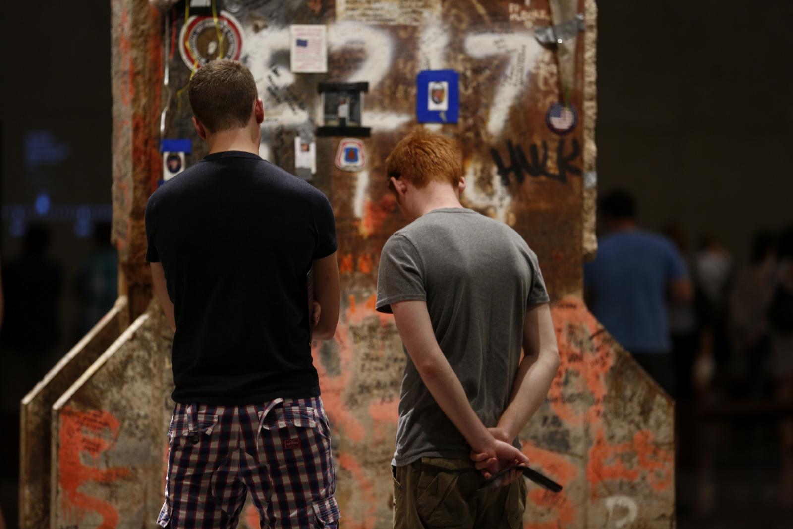 Two men face away as they look at the Last Column in Foundation Hall. The rusty column is covered in stickers, photos, handwritten messages, and other tributes, along with orange marking paint.