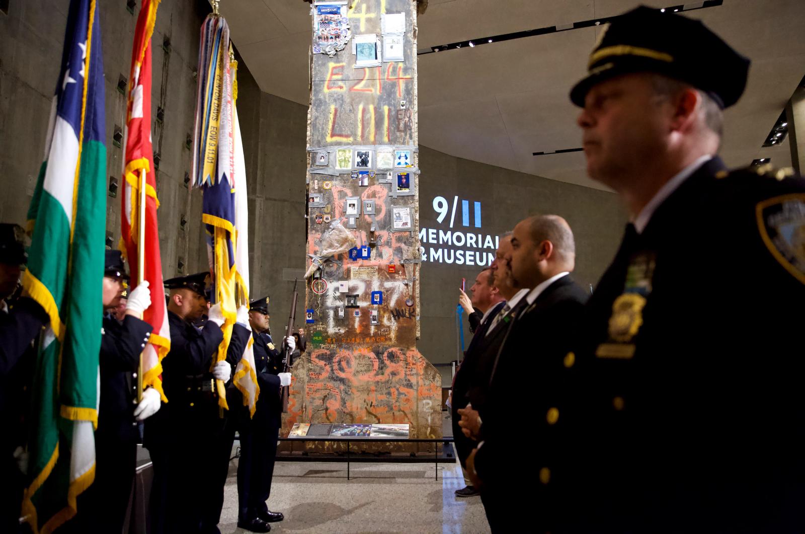 The Last Column towers over NYPD flag bearers standing to the left and men in suits and ties standing to the right. The Last Column, a fixture of Foundation Hall, is covered in photos and written tribute to rescue and recovery workers. 