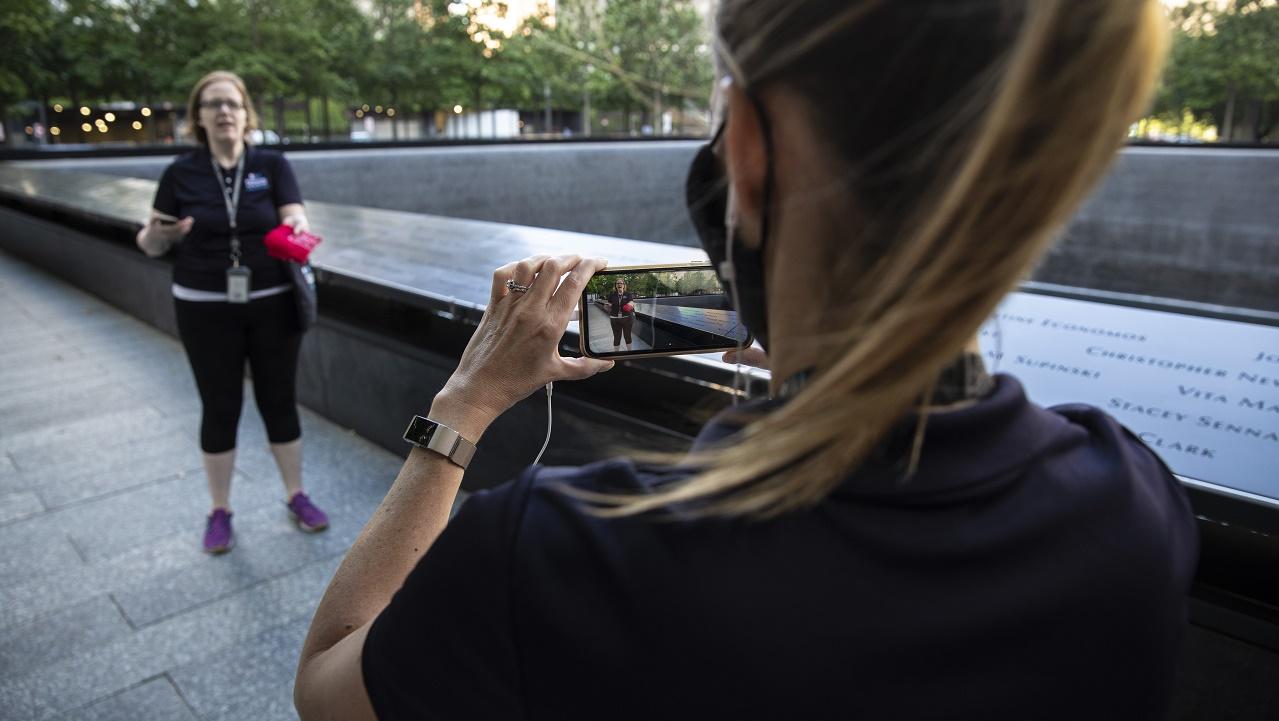A Museum employee in the foreground is shown from behind as she holds an i-phone up to record a guide leading a virtual experience on the 9/11 Memorial.