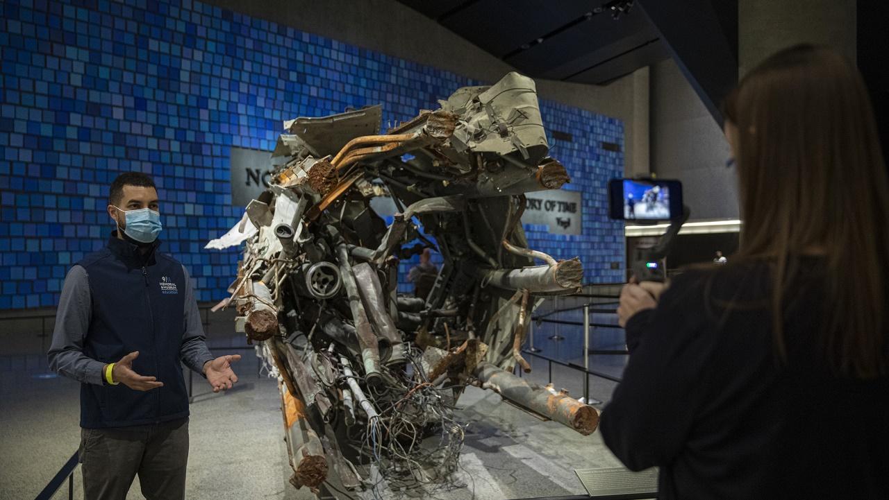 A Museum guide gestures as he stands in Memorial Hall near the 19.8-foot-long fragment recovered from the original 360-foot-tall radio and television antenna from atop the North Tower.  The guide is giving a virtual tour being filmed by another guide in the foreground of the image.