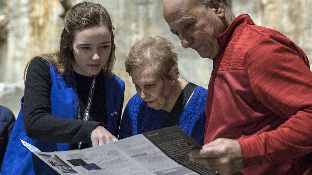 A Museum docent helps an elderly woman and man huddled to her left. The man and woman are holding a Museum guide. They look as the woman points to something on the guide with her right hand.