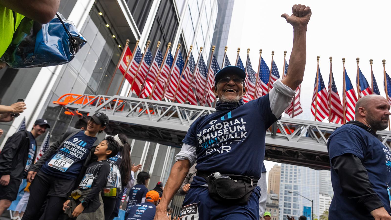 A person wearing navy blue shorts and a navy blue 5K t-shirt crosses the finish line, left arm raised in victory