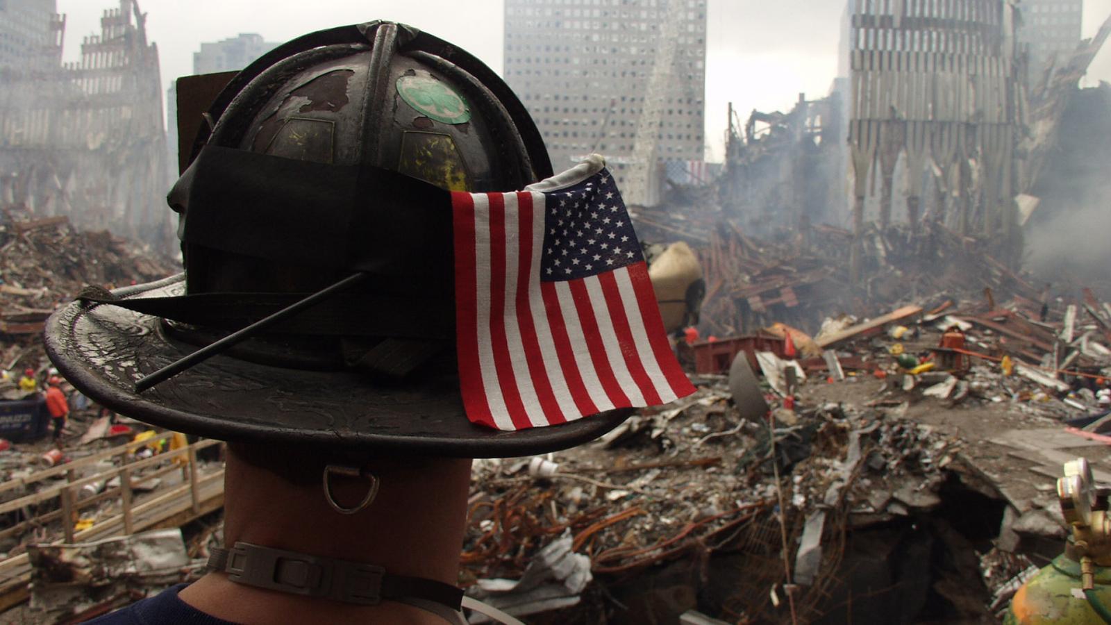Back view of a rescue worker in a hard hat with an American flag tucked in its rim, overlooking rubble at Ground Zero