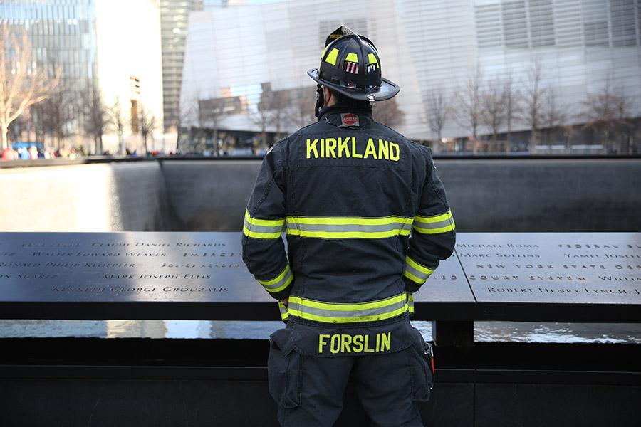 A firefighter pauses in reflection on 9/11 Memorial plaza as he looks out over a reflecting pool. The firefighter is wearing bunker gear that says “Kirkland” on the back.