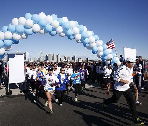 Dozens of people participating in the second annual 9/11 Memorial 5K Run/Walk jog past the start line, which consists of an arch made of blue and white balloons. The skyline of Jersey City can be seen in the distance on a sunny day.