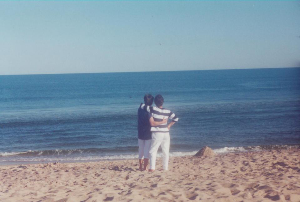 Alena Sesinova and Barbara Cattano embrace on a beach in the Hamptons. They are looking out at the ocean on a clear day.