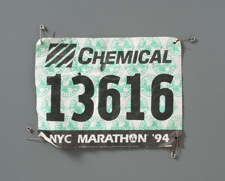 A marathon bib worn by NYPD Officer John Perry is displayed on a gray surface at the Museum. The word “chemical” is written on it, as are the words “NYC Marathon ‘94.” The number 13616 is at the center.