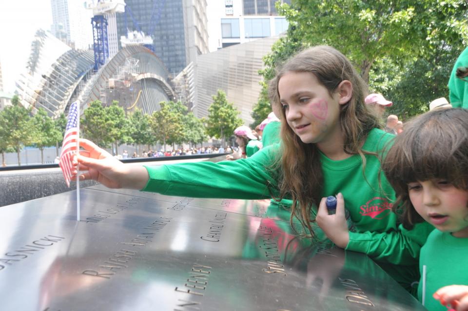 Several girls from Camp Simcha look at names on a bronze parapet at the 9/11 Memorial. A girl at center wearing a green sweatshirt is placing a small American flag on one of the names.
