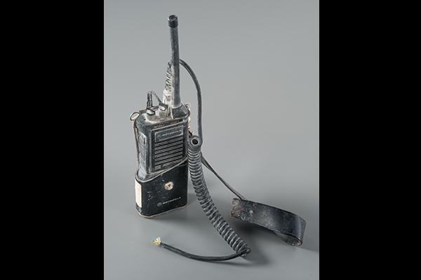 A radio used by FDNY fire chief Peter Ganci Jr. is displayed on a gray surface. The radio is damaged and covered in dust.