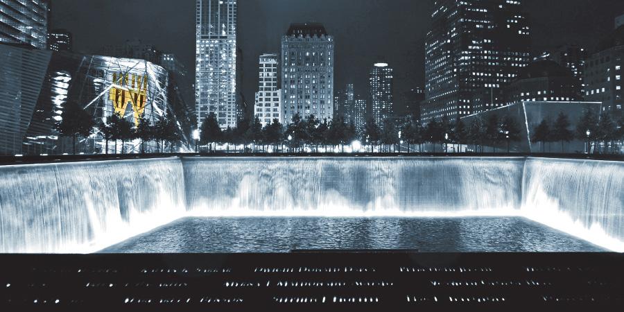 A monochrome image of the 9/11 Memorial shows one of the reflecting pools lit up at night, with surrounding buildings illuminated in the background. The Twin Towers’ steel, rust-colored tridents are the only colorized object in the photo. They are visible through the windows of the Museum Pavilion.